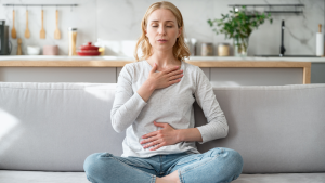 Woman sitting on a couch practicing deep diaphragmatic breathing exercises with one hand on her chest and one on her stomach to relieve anxiety.
