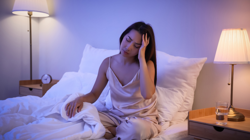 A frustrated woman sitting awake in bed at night, holding her head, illustrating the effects of insomnia and stress on sleep quality.