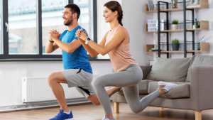 A smiling man and woman doing lunges in their living room, demonstrating a joyful and accessible way to practice mindful movement at home.