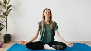 A woman sitting cross-legged on a blue yoga mat at home, practicing mindfulness meditation with her eyes closed to reduce stress.