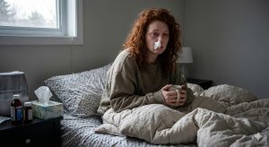 A miserable woman with red hair sitting up in bed with a tissue stuck in her nose, looking exhausted from constantly catching colds due to a weak immune system.
